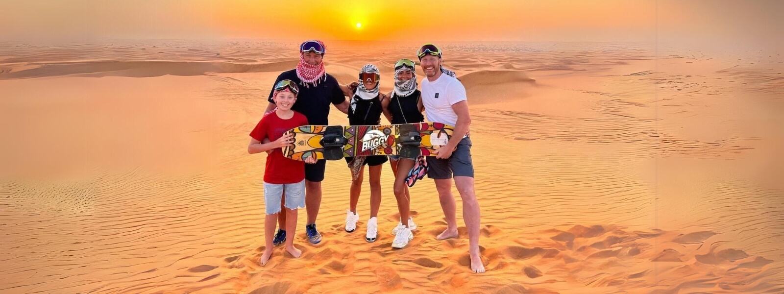 A family of five standing in the desert, holding a sandboard, enjoying a sunny day together.