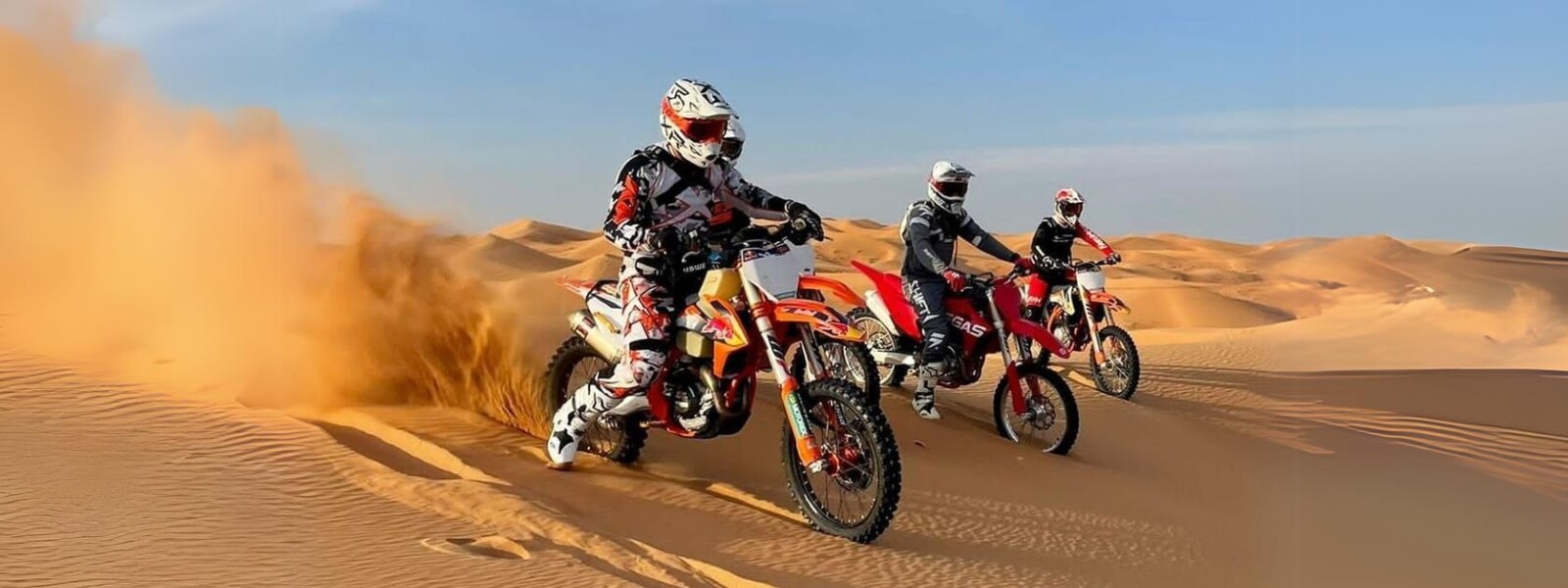 Three people riding dirt bikes in the desert, kicking up dust as they navigate the sandy terrain.