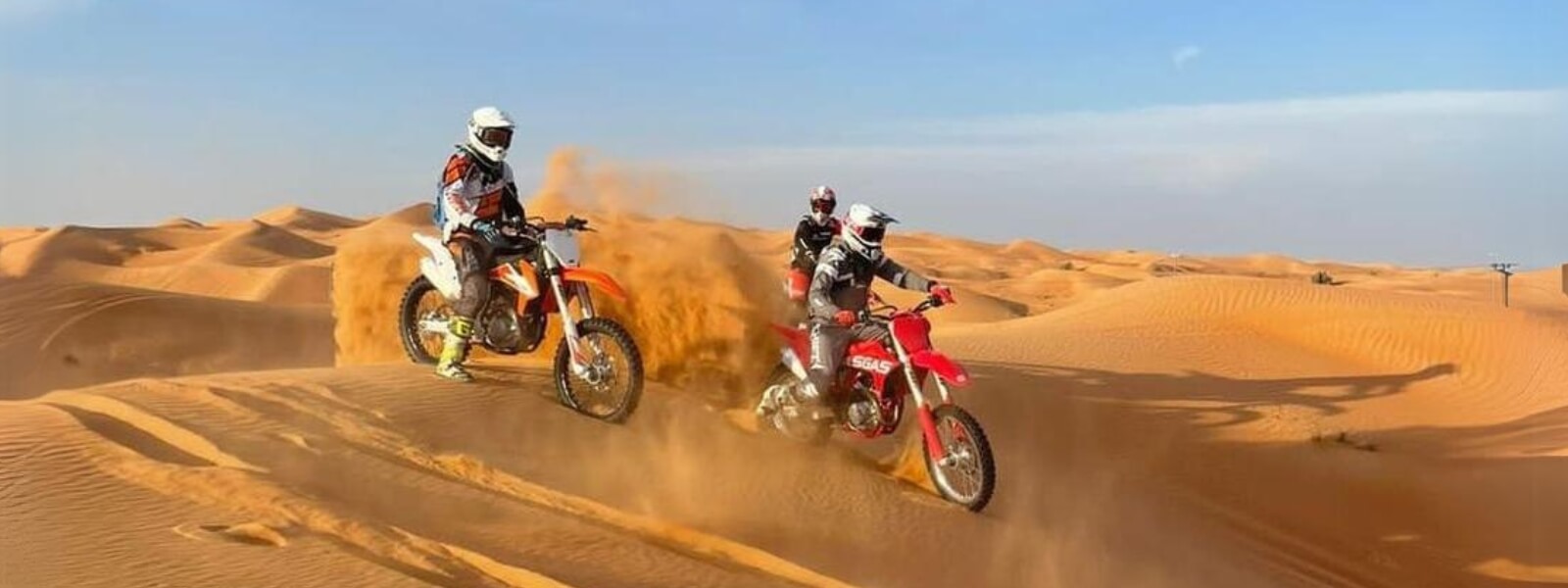 Two individuals ride dirt bikes in the Dubai desert under a clear blue sky.