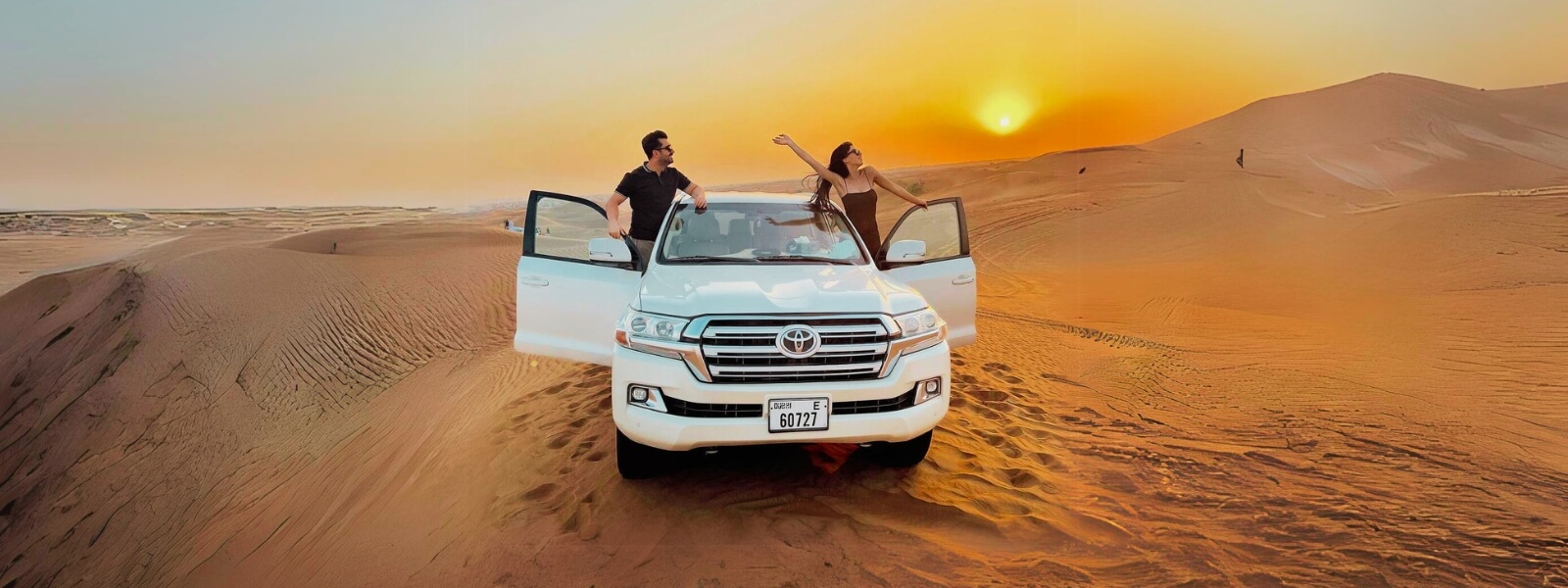 Two people riding on the back of a white SUV in the desert under a clear blue sky.