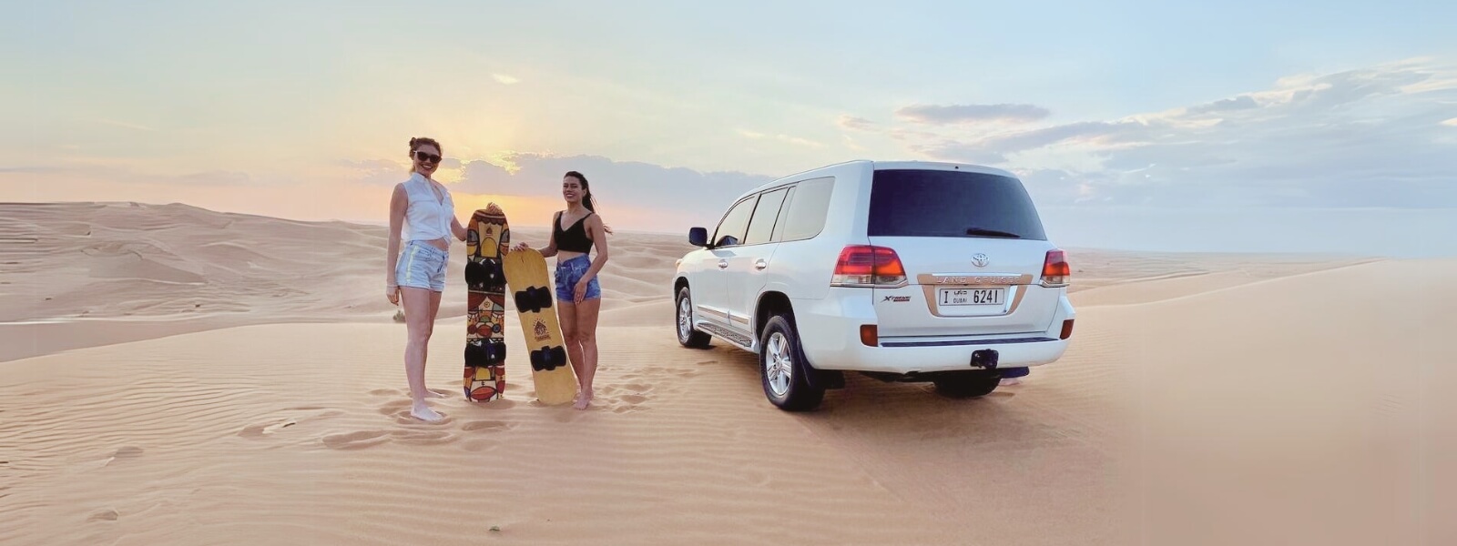 Two women standing near a white SUV in the desert, holding sandboards and enjoying an evening desert safari.
