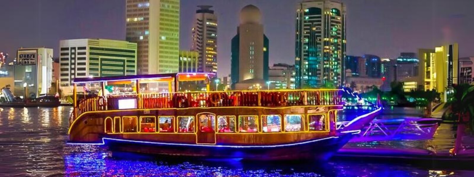A Dhow Cruise illuminated with lights sails in front of a city skyline at night.