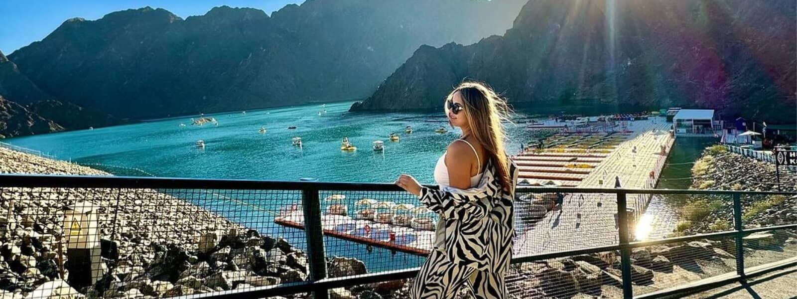 A woman standing on a railing, gazing at a serene lake surrounded by nature's beauty.